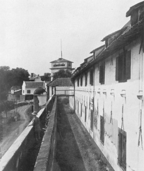 A scenery of ‘Oud Batavia’. On the right is an old VOC warehouse (now, used as the Jakarta Maritime Museum), in the middle is the part of a city wall with a sentry box, in the distance is the Watchtower (Vries, J.J. De (ed.), Jaarboek van Batavia en omstreken 1927, Kolff, Batavia 1927)